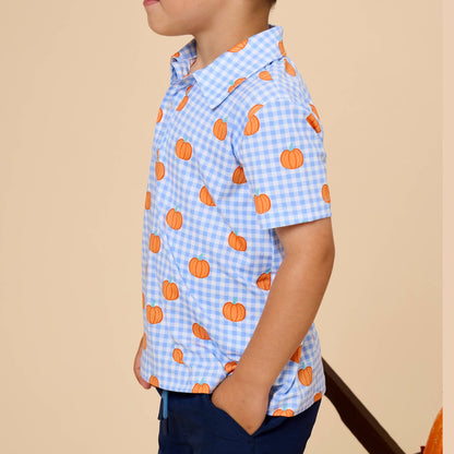 A young boy in profile wearing a light blue and white gingham short-sleeve button-up shirt with an orange pumpkin print, paired with navy pants, set against a solid beige background.
