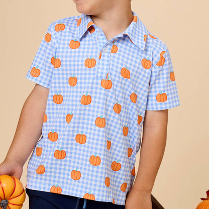 A young boy smiling and looking to the side, wearing a light blue and white gingham short-sleeve button-up shirt with an orange pumpkin print, paired with navy pants. He is holding a small orange pumpkin against a solid beige background.