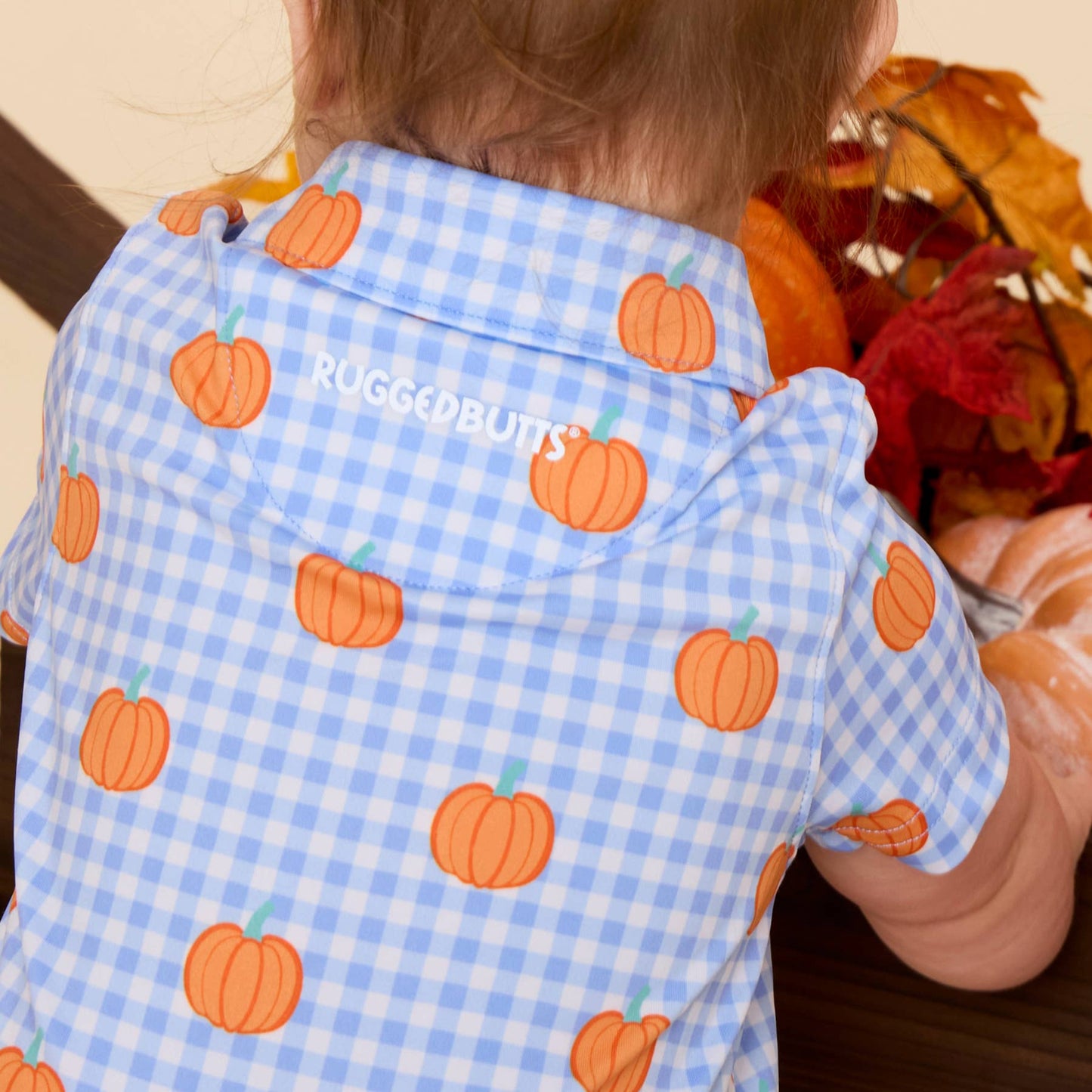 A baby viewed from behind wearing a light blue and white gingham short-sleeve shirt with an orange pumpkin print. The brand name 'RUGGEDBUTS' is printed in white on the back of the shirt.