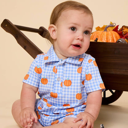 A baby sitting on a beige studio floor wearing a light blue and white gingham short-sleeve romper with an orange pumpkin print. A dark wooden wheelbarrow filled with autumn decorations is behind the baby, and two small decorative pumpkins are in front.