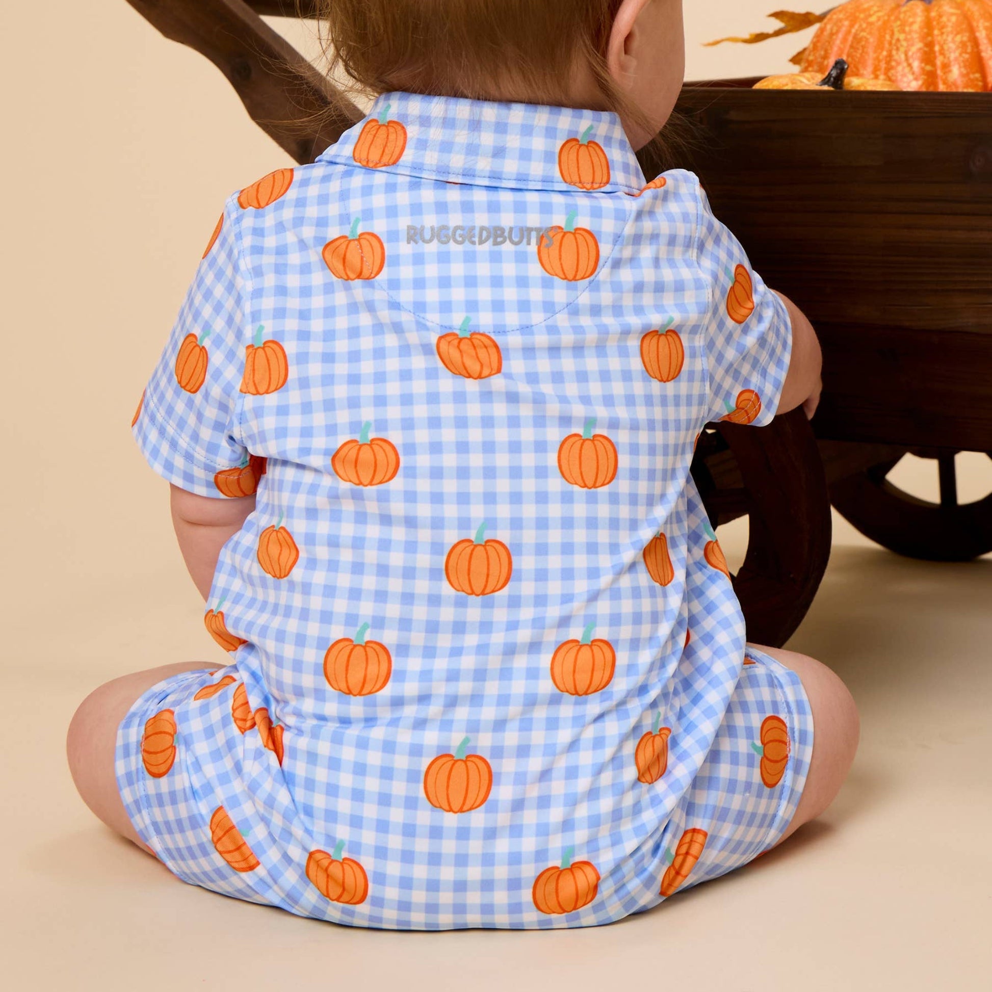 A baby seen from behind wearing a light blue and white gingham short-sleeve romper with an orange pumpkin print, sitting in front of a wooden wheelbarrow on a beige background.