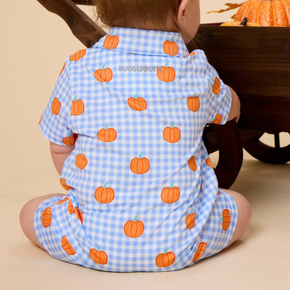 A baby seen from behind wearing a light blue and white gingham short-sleeve romper with an orange pumpkin print, sitting in front of a wooden wheelbarrow on a beige background.