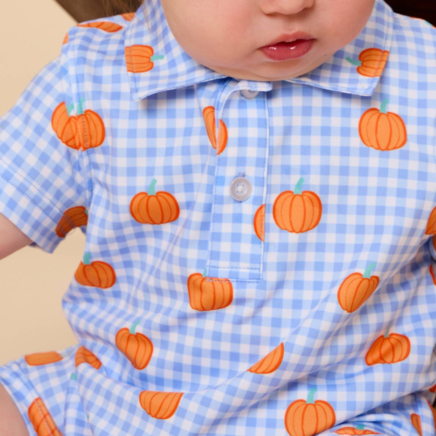 A close-up of a baby wearing a light blue and white gingham short-sleeve polo romper with an orange pumpkin print.