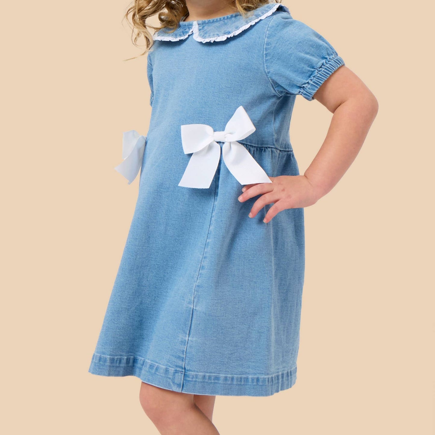 A young girl with blonde hair wearing a light blue denim dress with white ruffle trim on the collar and a large white bow at the waist, posing against a solid beige background.