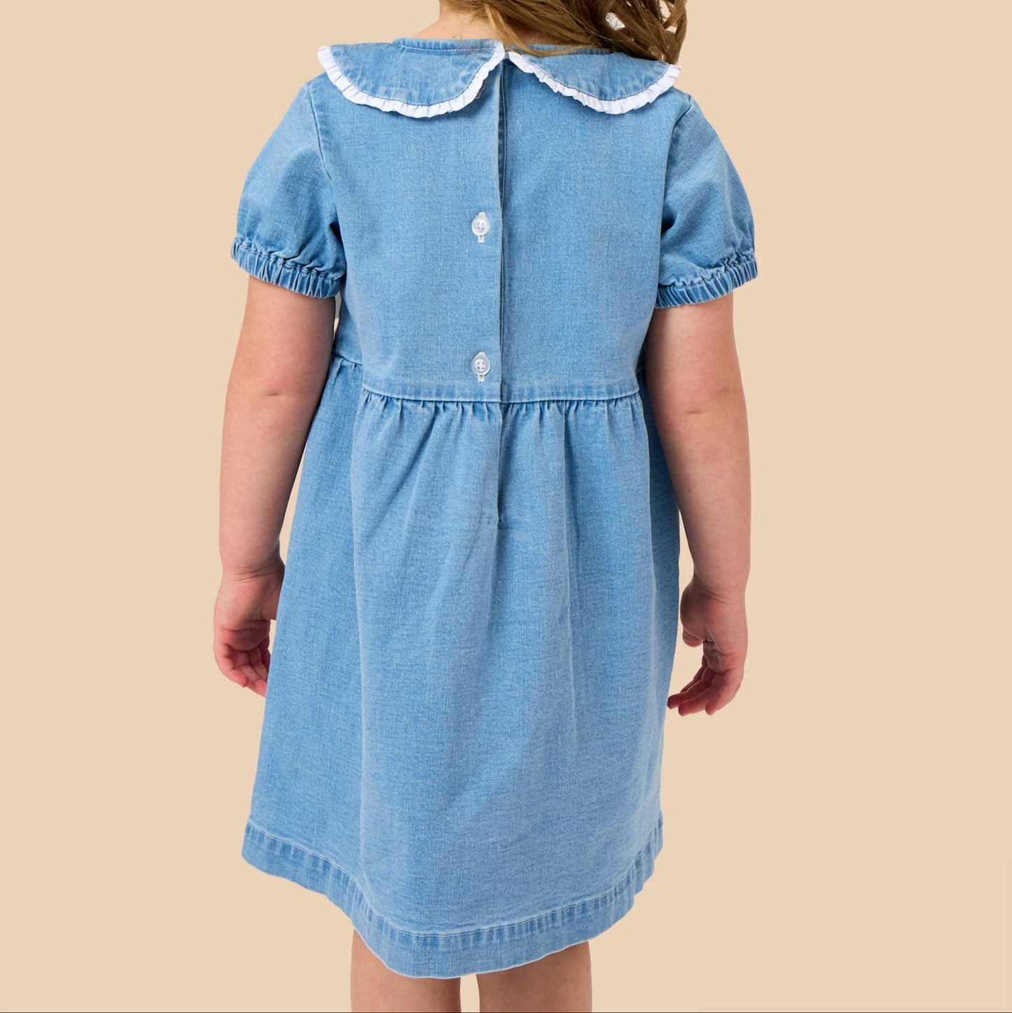 A young girl viewed from behind wearing a light blue denim dress with a white ruffled Peter Pan collar and white buttons. She has a large white bow in her hair, set against a solid beige background.