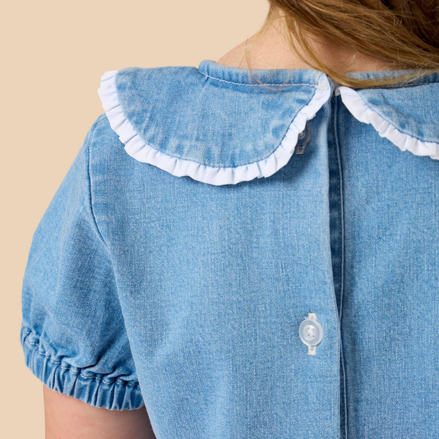 Close-up back view of a child wearing a light blue denim dress with a white ruffled Peter Pan collar and white buttons, against a solid beige background.