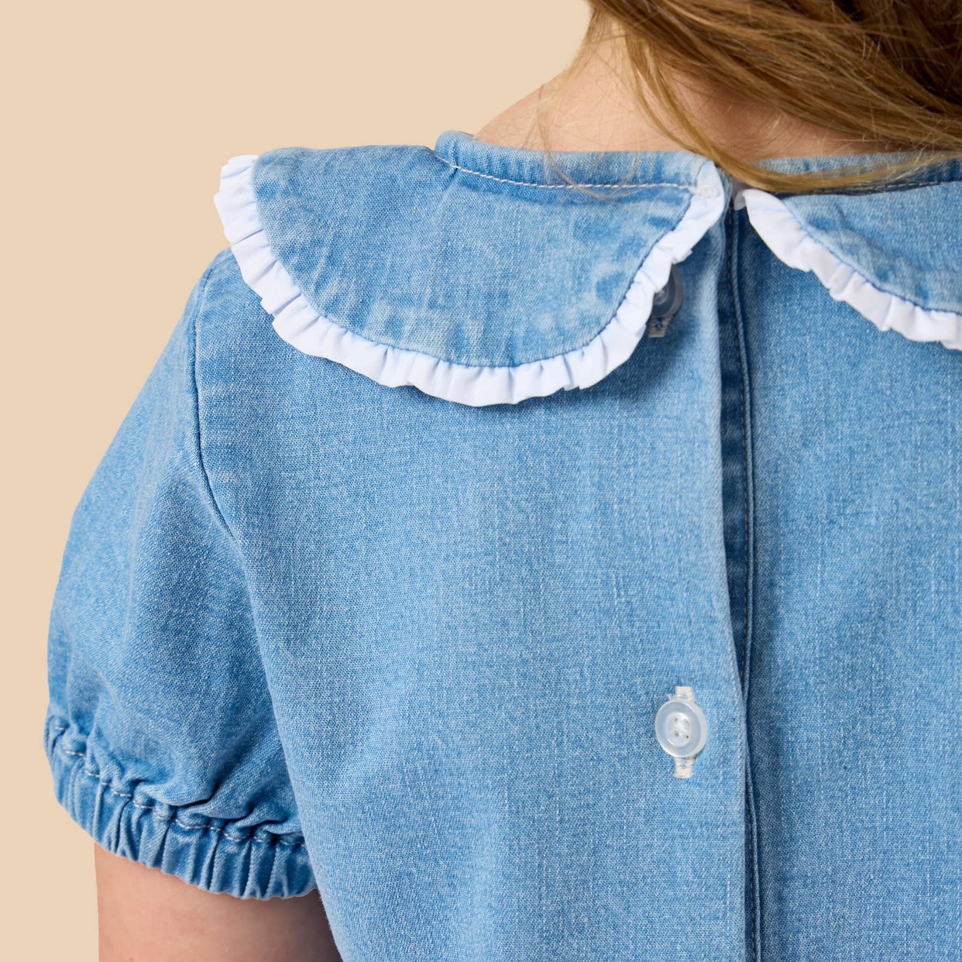 Close-up back view of a child wearing a light blue denim dress with a white ruffled Peter Pan collar and white buttons, against a solid beige background.