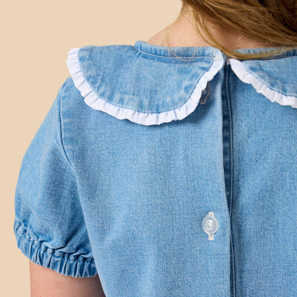 Close-up back view of a child wearing a light blue denim dress with a white ruffled Peter Pan collar and white buttons, against a solid beige background.