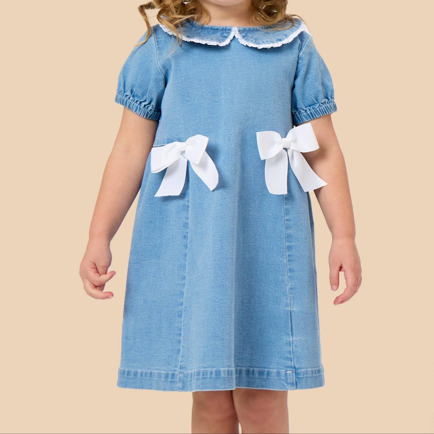 A young girl wearing a light blue denim dress with white accents, including a ruffled collar and two large white bows on the front, standing against a solid beige background.