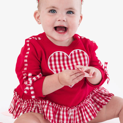 Baby wearing a red outfit with a checkered heart design and a large red bow.
