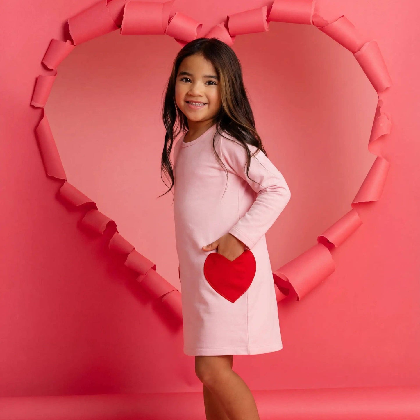 Young girl in a pink dress with a red heart design, standing in front of a heart-shaped frame on a pink background.