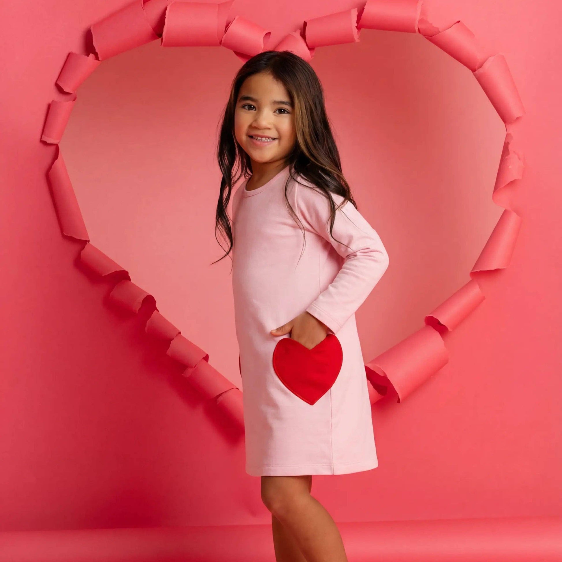 Young girl in a pink dress with a red heart design, standing in front of a heart-shaped frame on a pink background.