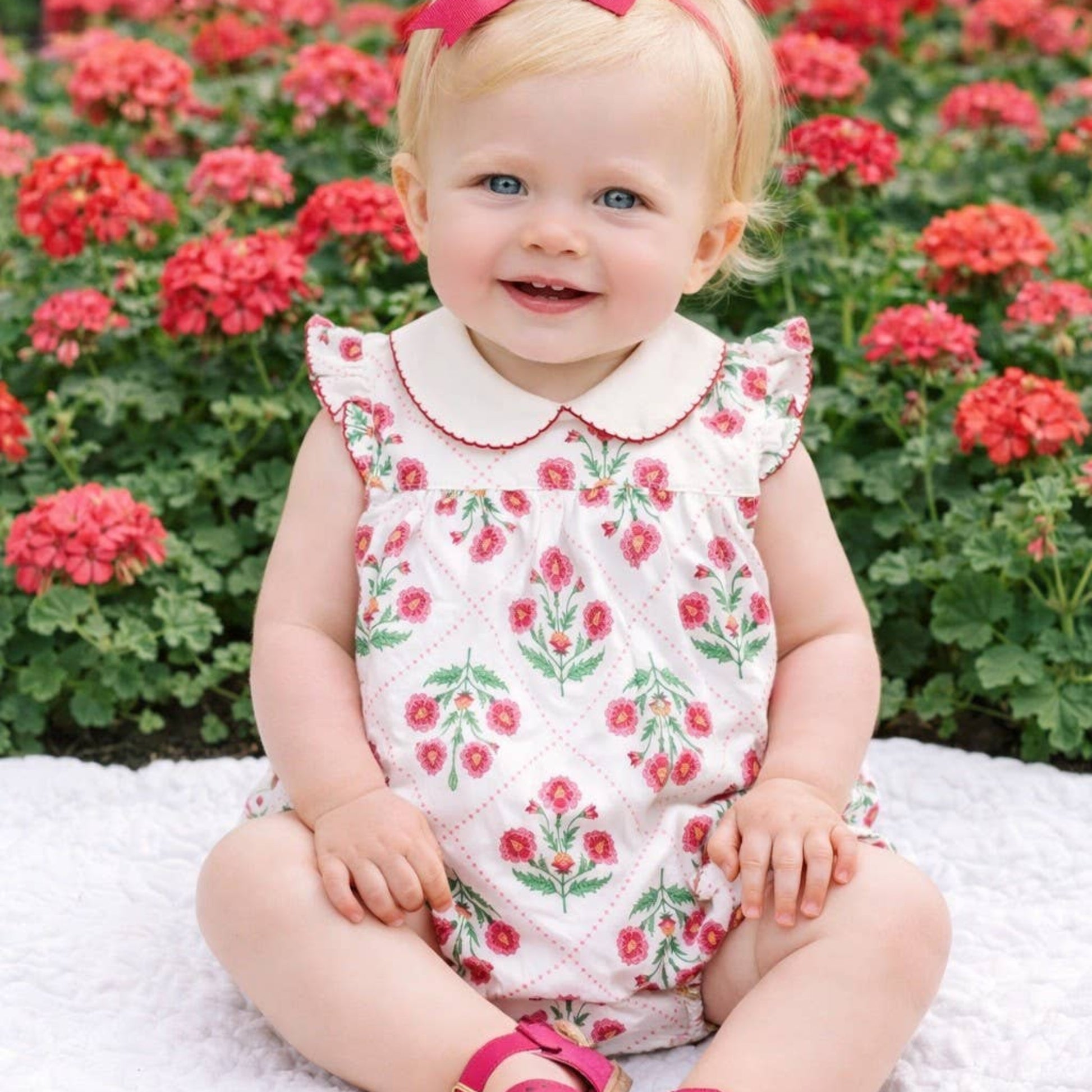 Child wearing a floral dress and pink sandals sitting in front of red flowers.