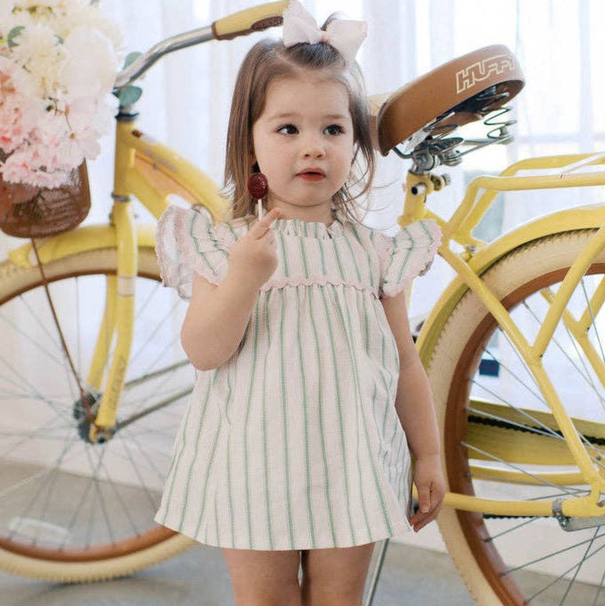 Young girl in a striped dress standing next to a yellow bicycle with a basket of flowers.