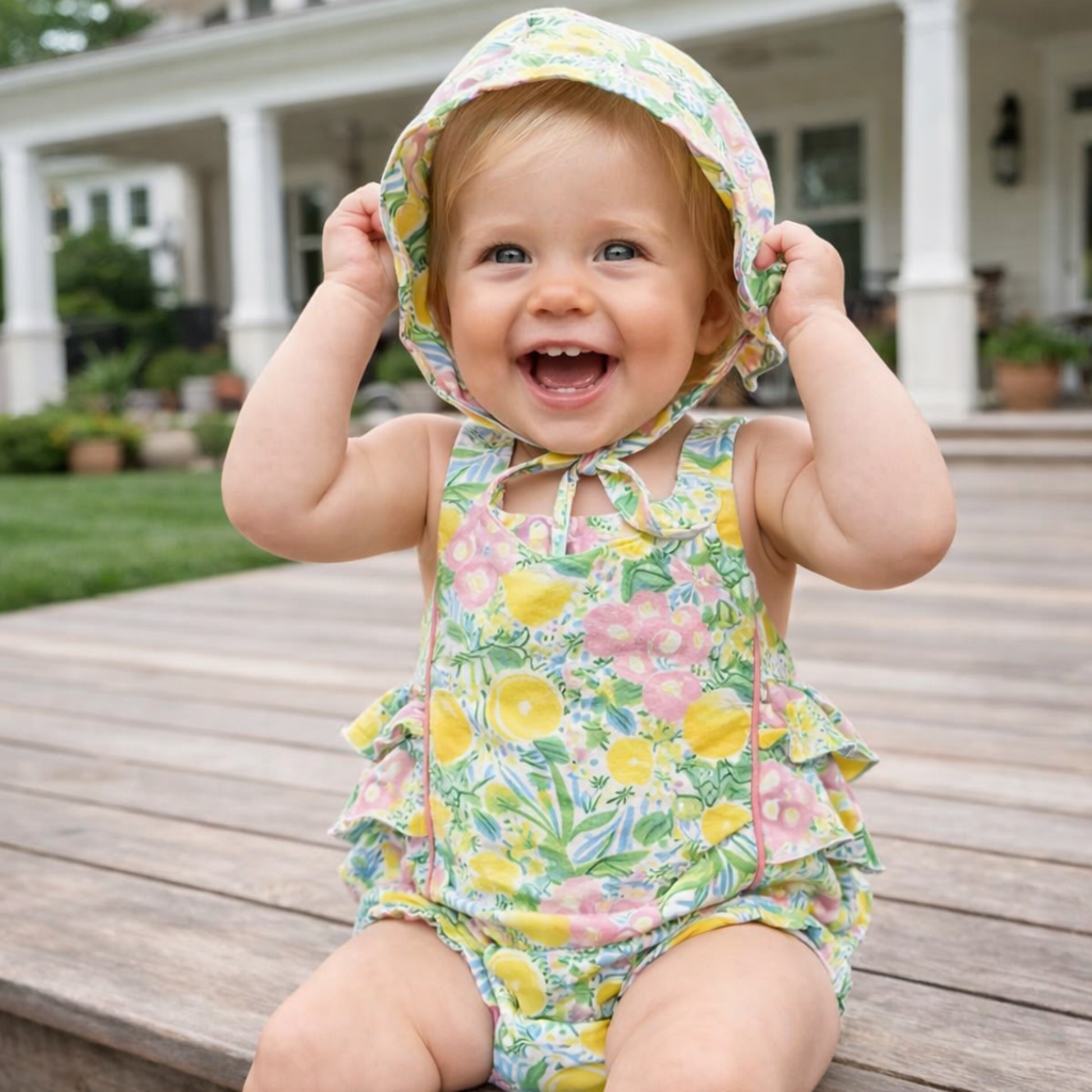Child wearing a floral sunsuit with a matching bucket hat, sitting on wooden steps outdoors.