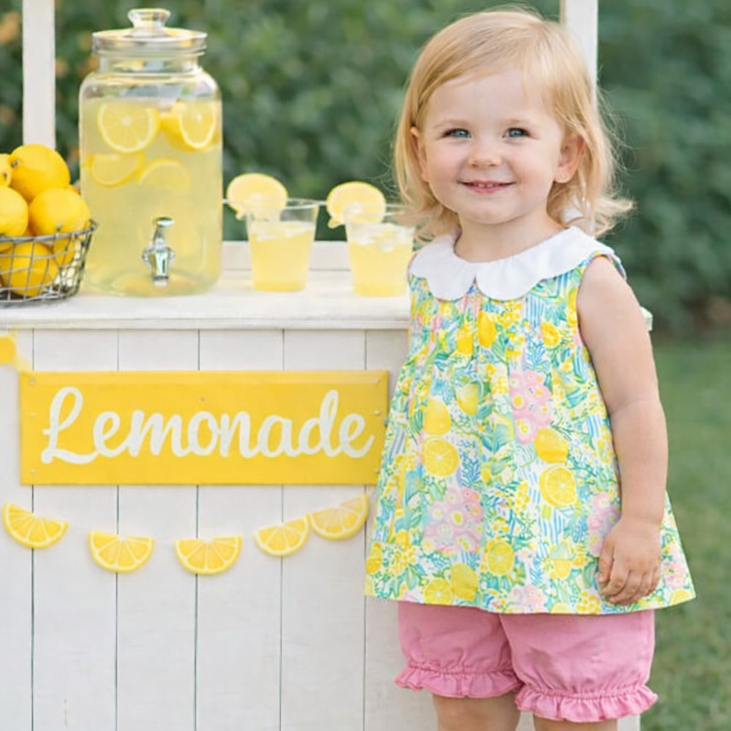 Child standing next to a lemonade stand with lemonade and lemons.