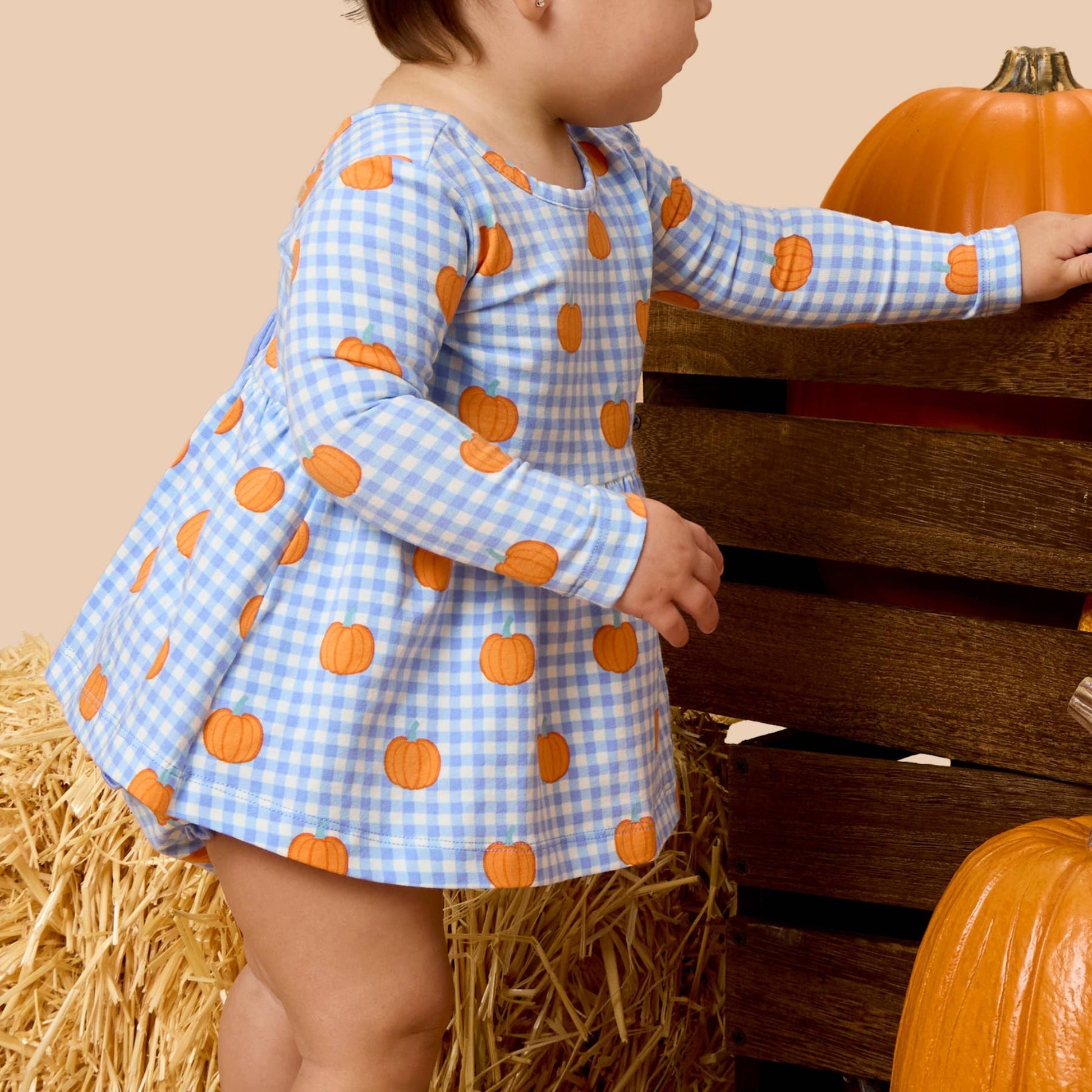 A toddler wearing a light blue and white gingham long-sleeve dress with orange pumpkin prints, standing next to a wooden crate with pumpkins on a beige background.