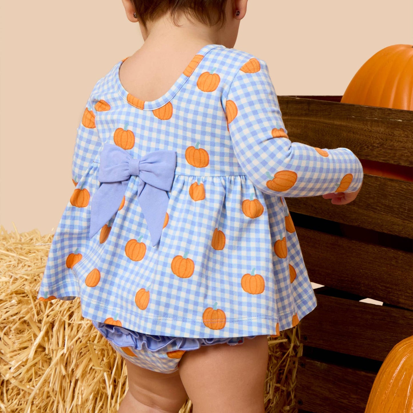 A toddler viewed from behind wearing a light blue and white gingham long-sleeve top with orange pumpkin prints and a large light blue bow on the back, standing beside hay bales and pumpkins.