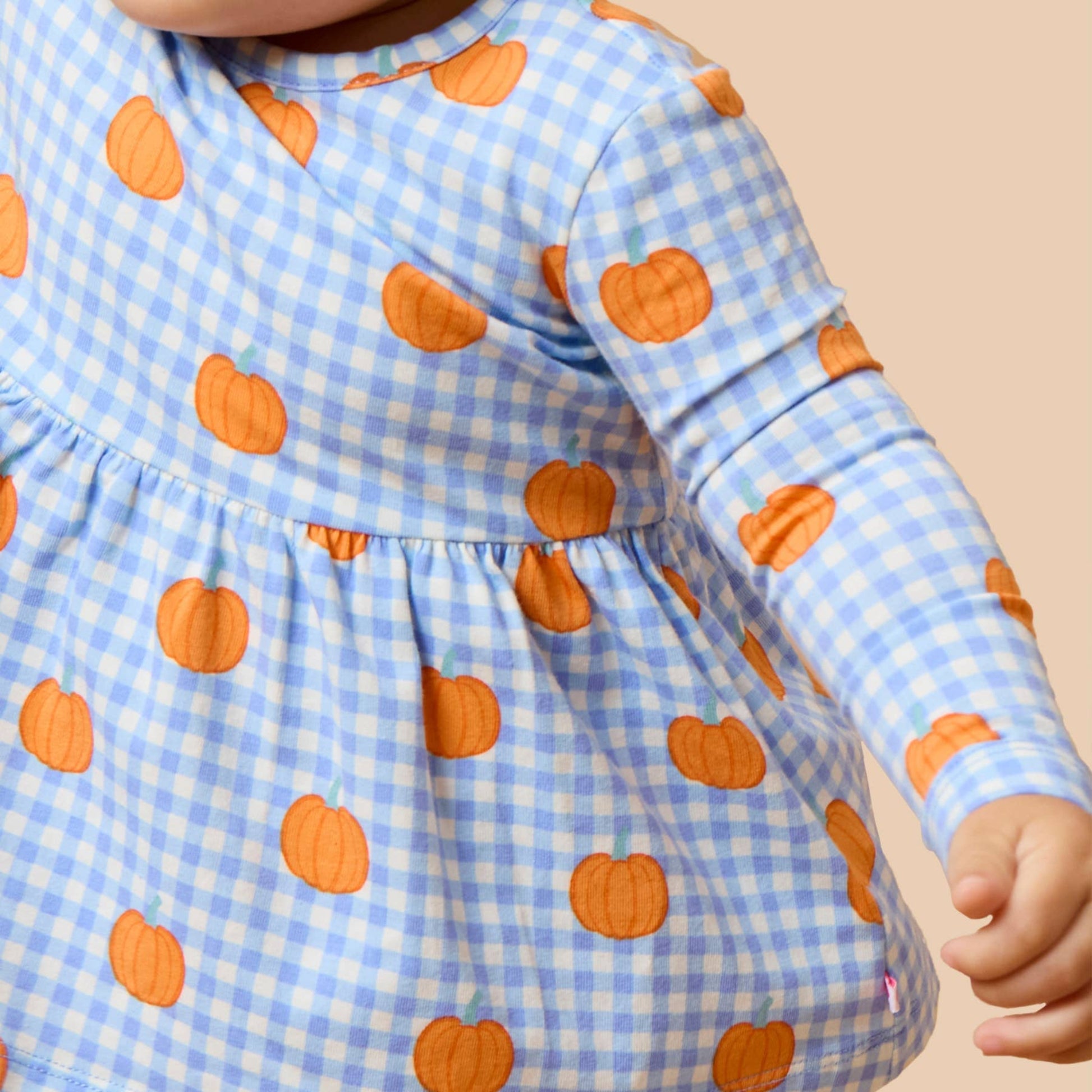 A close-up of a baby wearing a light blue and white gingham long-sleeve dress with an orange pumpkin print, set against a solid beige background.