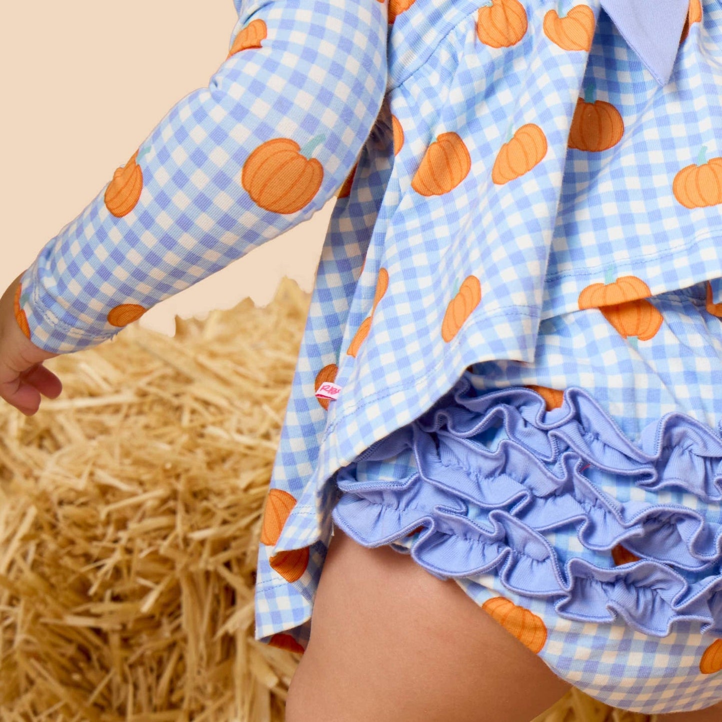 A close-up of a child wearing a light blue and white gingham romper with orange pumpkin prints, featuring a ruffled bloomers leg and a matching bow at the back, posed next to a bale of straw.