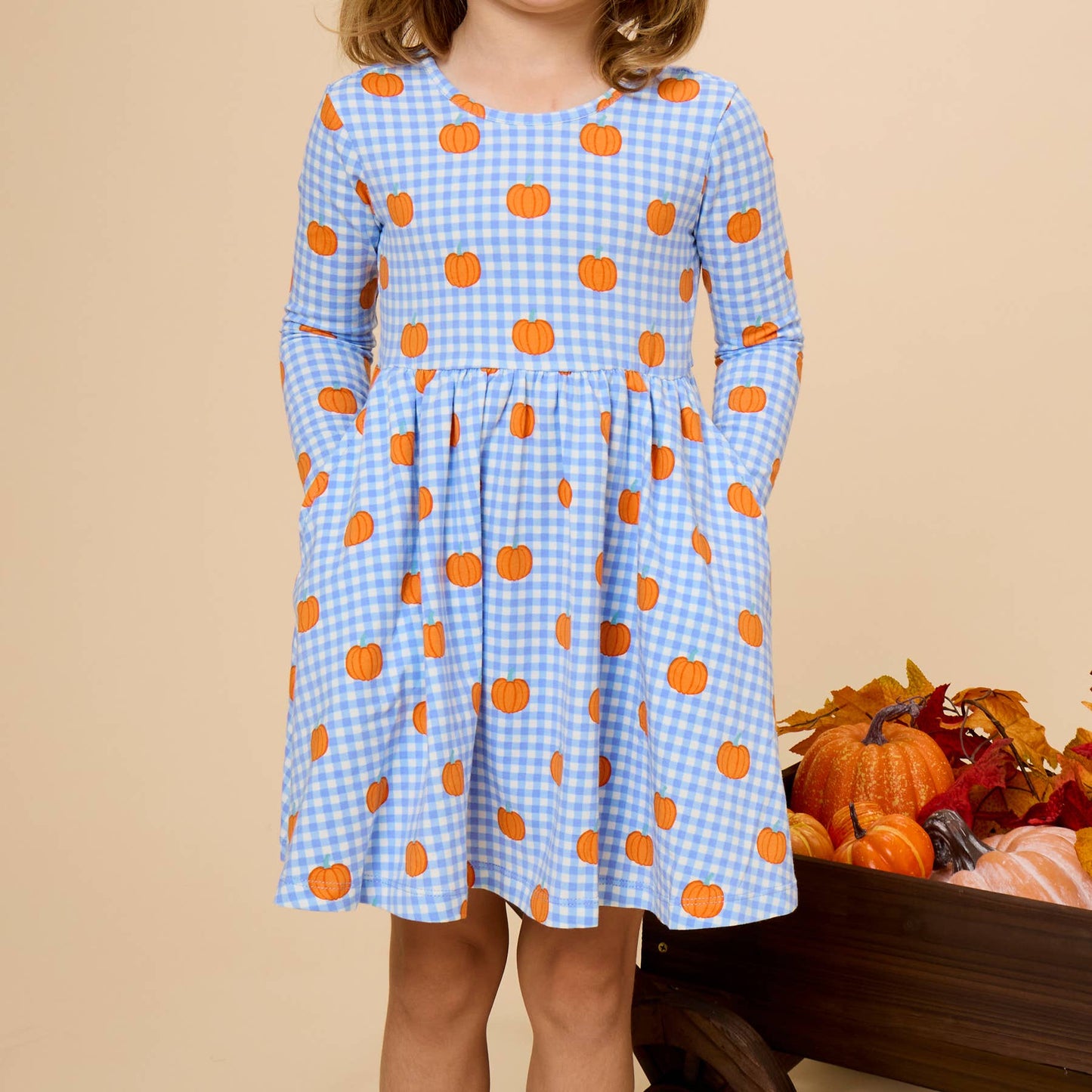 A young girl with brown hair wearing a light blue and white gingham dress with orange pumpkin prints, standing in front of a beige background with a decorative autumn pumpkin basket.