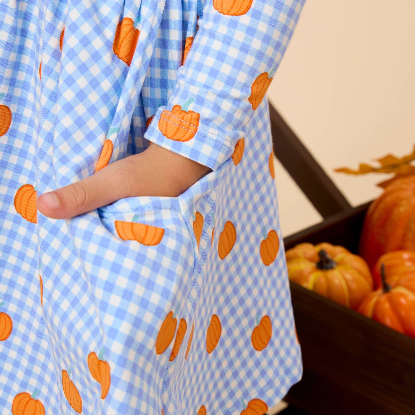 A close-up of a child wearing a light blue and white gingham dress with orange pumpkin prints, featuring a side pocket with the child's hand inside.