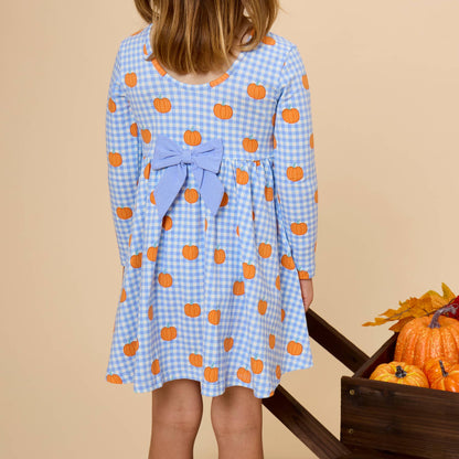 A young girl viewed from behind wearing a light blue and white gingham long-sleeve dress with orange pumpkin prints and a matching blue bow at the back waist. She is standing next to a wooden crate of pumpkins against a solid beige background.