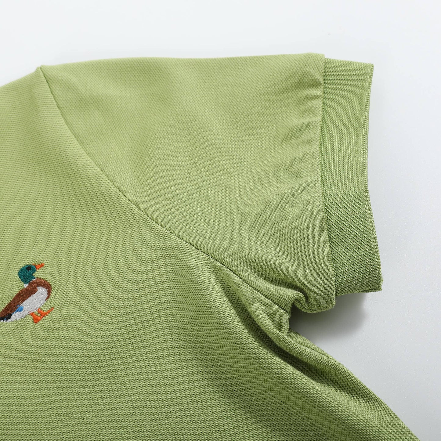A close-up of a light olive green polo shirt showing the collar, sleeve cuff, and a small embroidered mallard duck logo on the chest, set against a plain white background.