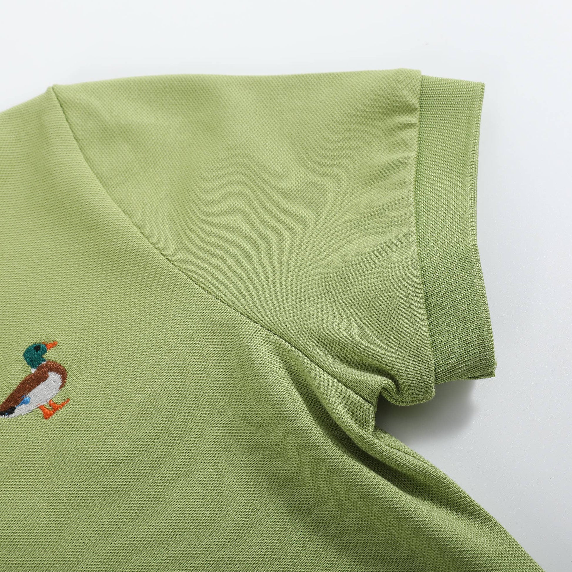 A close-up of a light olive green polo shirt showing the collar, sleeve cuff, and a small embroidered mallard duck logo on the chest, set against a plain white background.