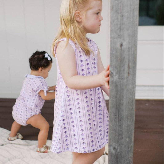 A young blonde girl wearing a sleeveless lavender and white patterned dress with a white bow in her hair stands next to a wooden post on a porch, while a toddler in a matching outfit crouches in the background.