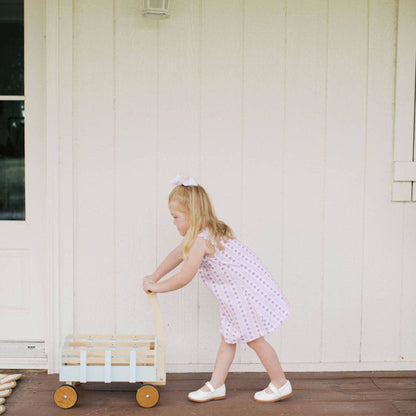 A young girl with blonde hair in a white bow pushes a small wooden toy wagon on a brown wooden porch in front of a white house.
