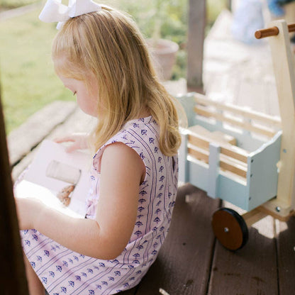 A young girl with blonde hair and a white bow sits on a wooden porch, wearing a light pink and white patterned sleeveless dress. A light blue wooden toy cart is visible next to her in the background.