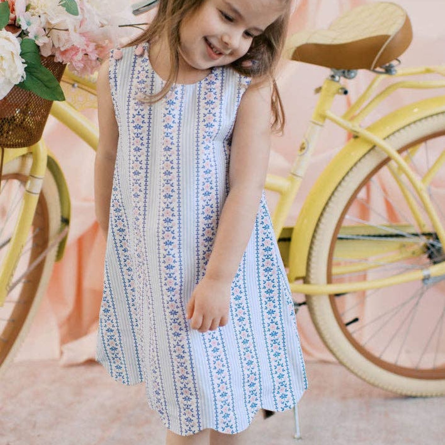Young girl in a patterned dress standing next to a bicycle with a floral arrangement.