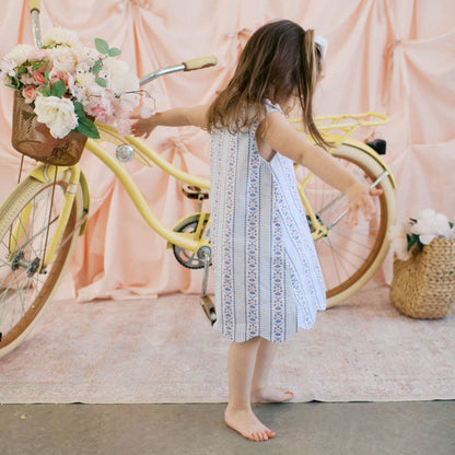 Young girl in a white dress standing next to a yellow bicycle with a floral basket against a pink curtain backdrop.