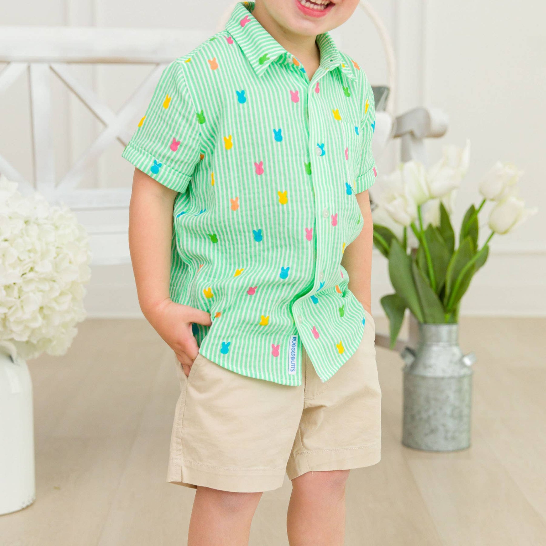 Child wearing a green patterned shirt with colorful squares and beige shorts, standing indoors.