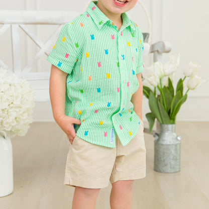 Child wearing a green patterned shirt with colorful squares and beige shorts, standing indoors.